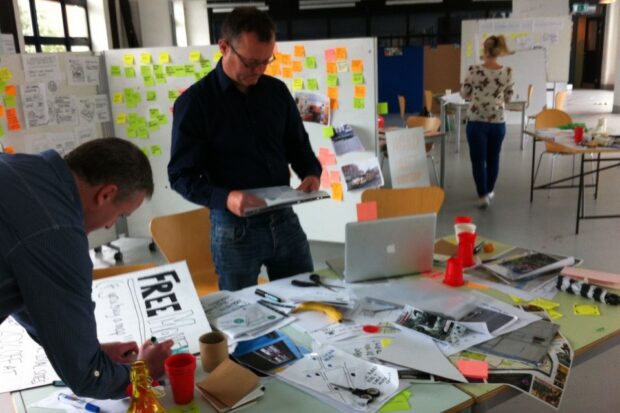 Picture of two men considering ideas outputs, one standing, the other leaning over a table