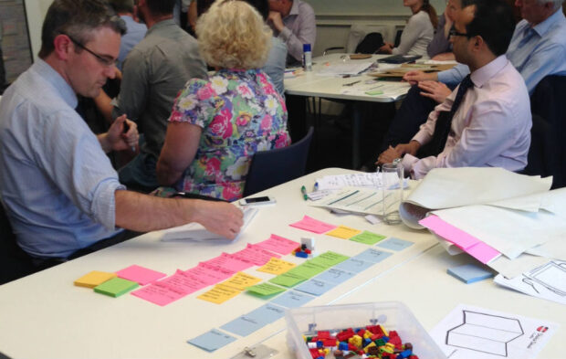 A man is sitting at a table, concentrating on writing on post-it notes that are lined up by colour. On a table behind him a group of people are listening to a speaker. There is a box of lego on the table.