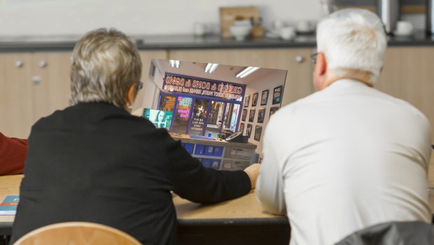 Two people are shot from behind, sitting next to one another. In between them they are holding and considering a poster of the image from within a shop, looking out through the shopfront.